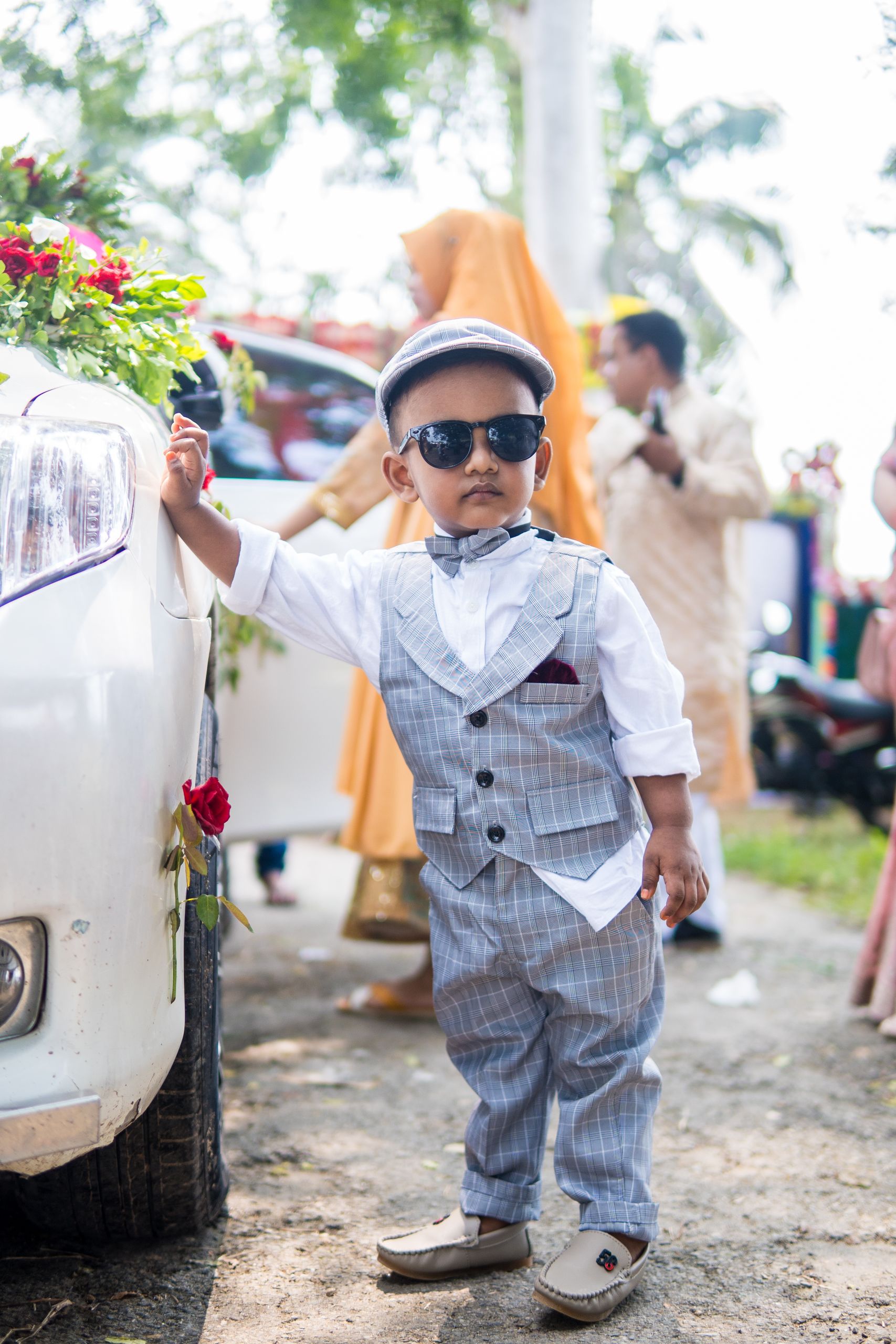 A fashionable young boy in a suit poses by a decorated car during a sunny outdoor wedding.