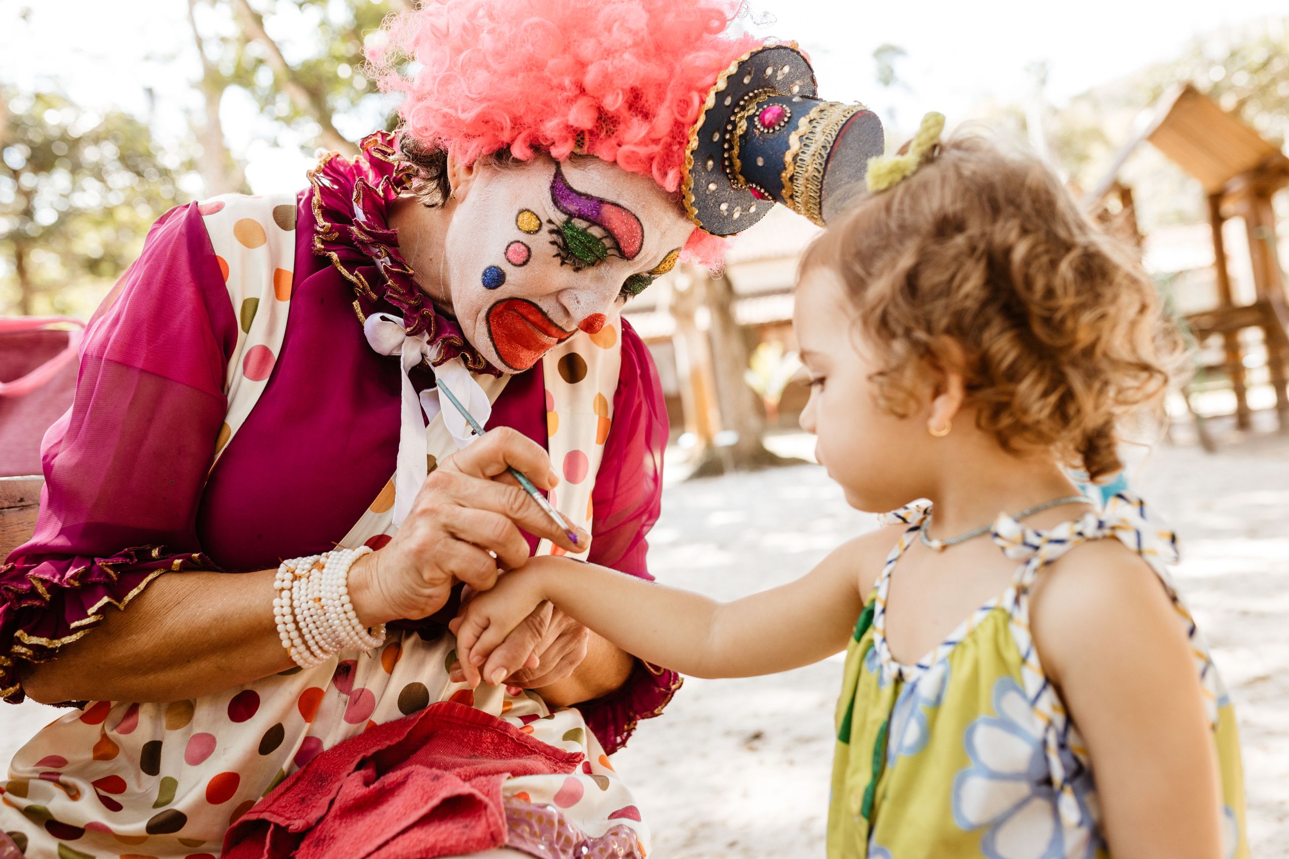 A vibrant clown paints a child's hand at a lively outdoor party, creating a joyful atmosphere.
