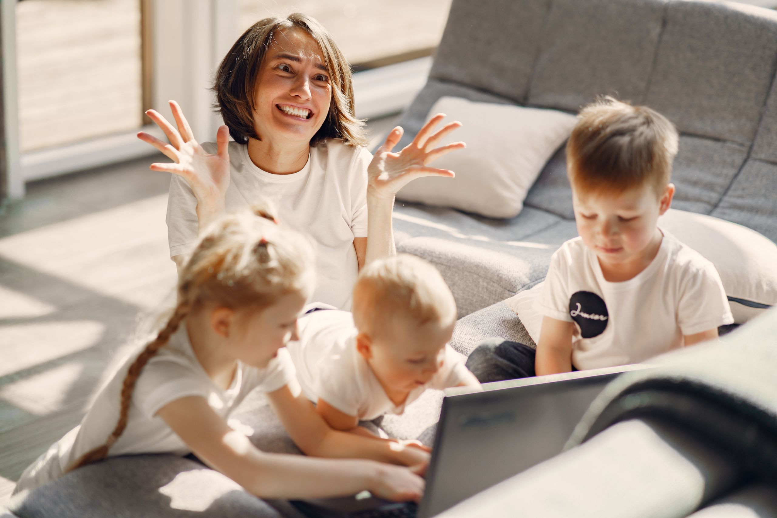 From above of woman making grimace while stressing about looking after little kids playing on laptop together on sofa in living room