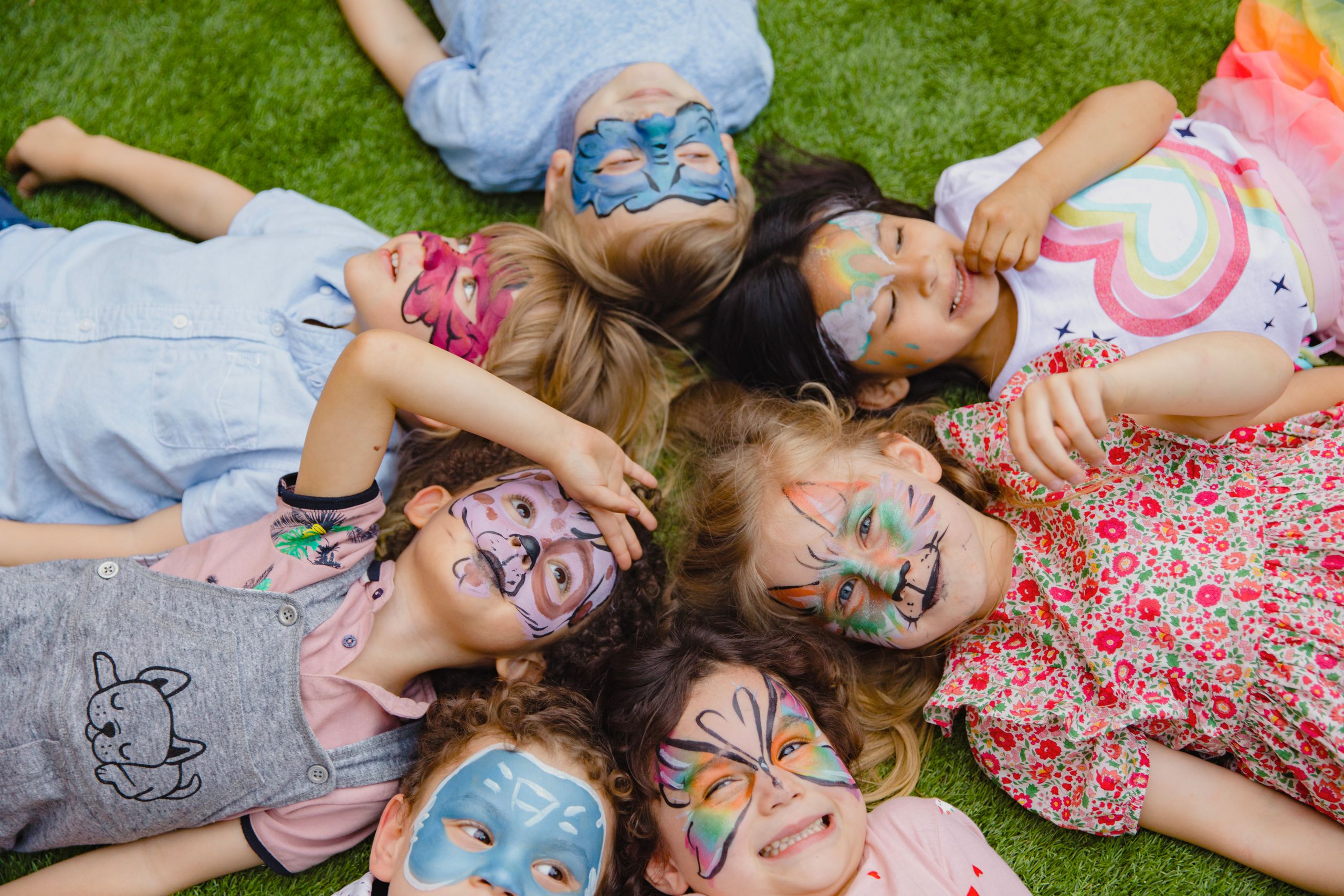 Group of children enjoying an outdoor party with colorful face paint, lying on grass.
