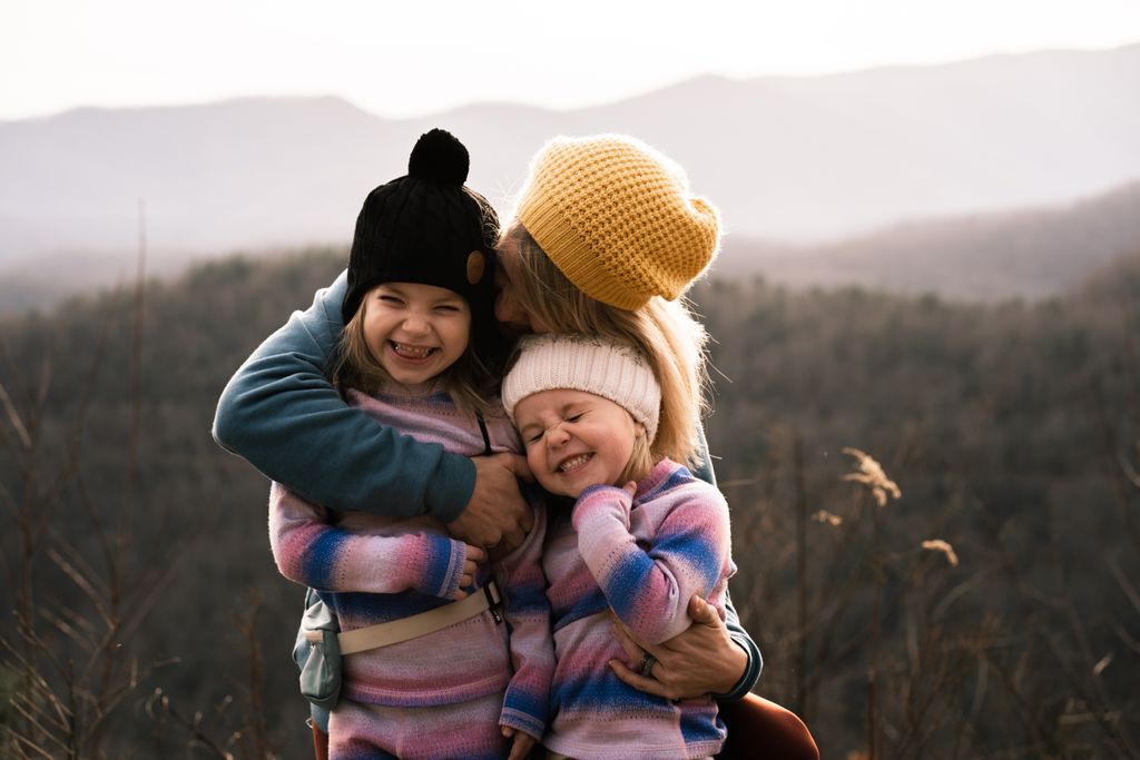 Mother embracing daughters with smiles in Asheville's scenic fall landscape.