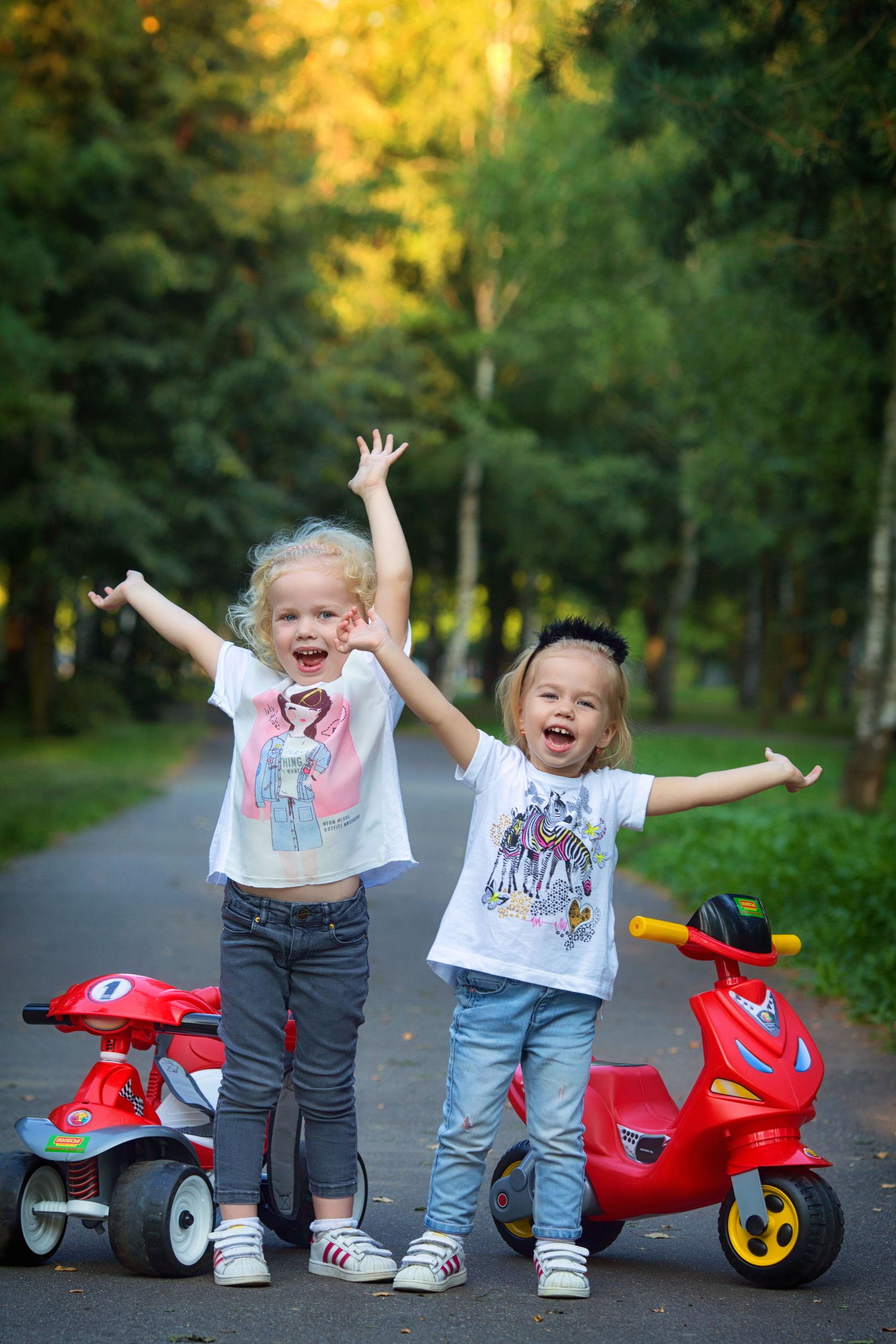 Two smiling children enjoy a sunny day outdoors with their toy motorbikes, surrounded by trees.